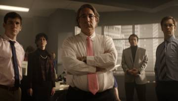 An actor portraying Bernie Madoff has his arms crossed, standing in front of his colleagues in an office building.