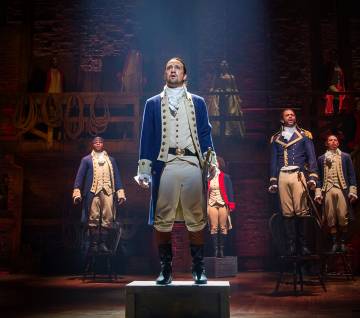Lin Manuel Miranda and the cast of Hamilton standing on a Broadway stage. 