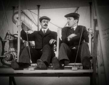 Two actors portraying the Wright brothers argue as their flying a very old-fashioned style airplane. 