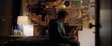 A teenage boy is sitting on his bedroom looking at his cell phone.  The wall behind him is covered in music and space posters.  