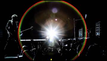 Bono is standing on a stage with a guitar in front of crowd of people and the flash from a camera behind Bono creates a red and green circle around the stage.