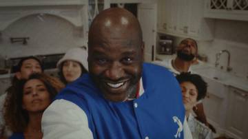 A selfie taken by Shaquille O'Neal who is standing in a white kitchen, smiling, looking directly into the camera. Five other people are standing behind him also looking into camera.